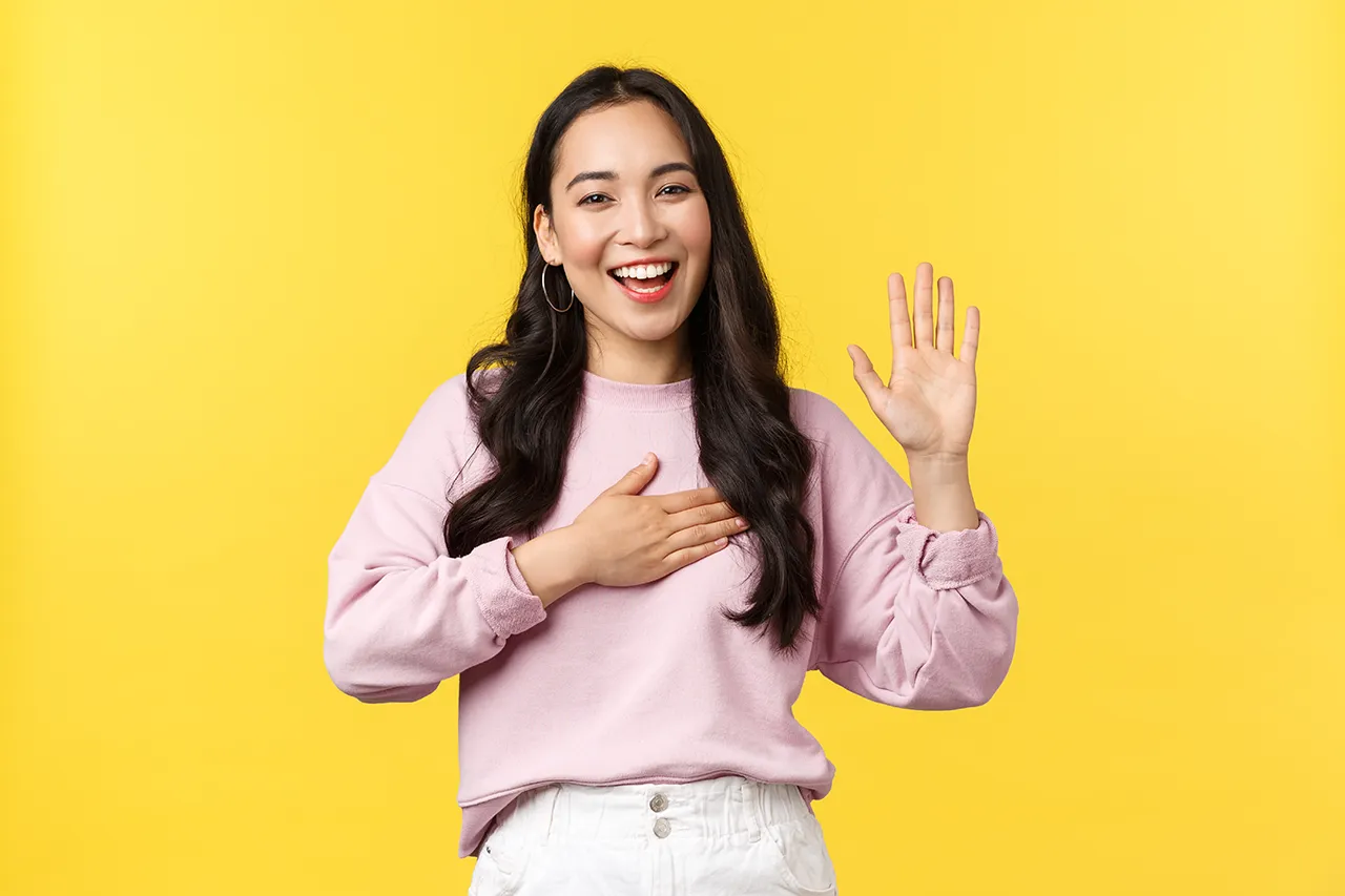 Lachende jonge vrouw met lichtroze trui groet met open hand en hand op haar hart tegen gele achtergrond