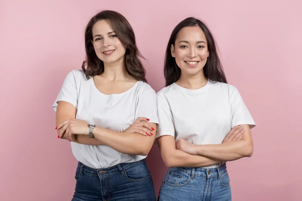 Twee jonge vrouwen staan tegen roze achtergrond en kijken zelfverzekerd de camera in met de armen over elkaar heen