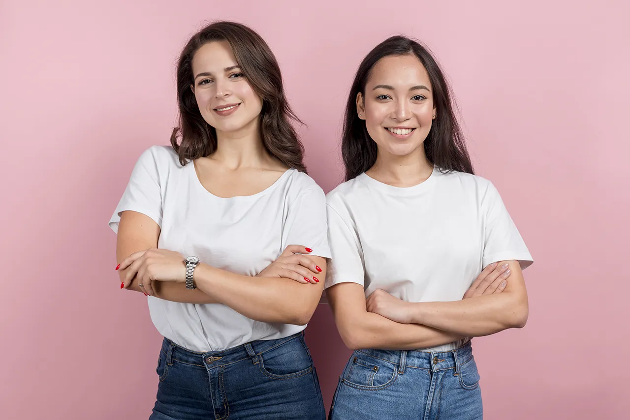 Twee jonge vrouwen staan tegen roze achtergrond en kijken zelfverzekerd de camera in met de armen over elkaar heen