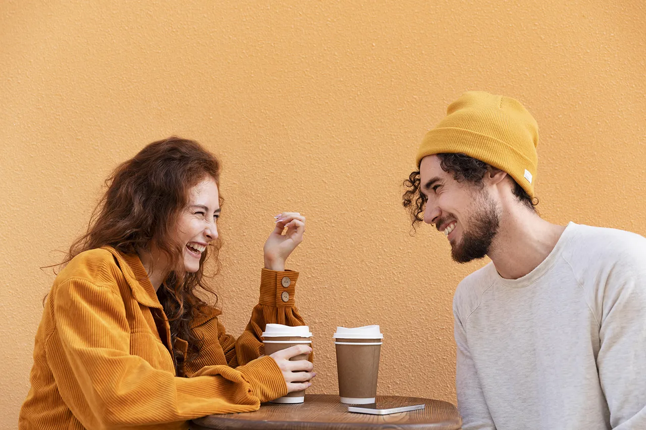 Vrouw met oranje trui en man met gele muts lachen terwijl ze een kop koffie drinken uit kartonnen beker tegen oranje achtergrond