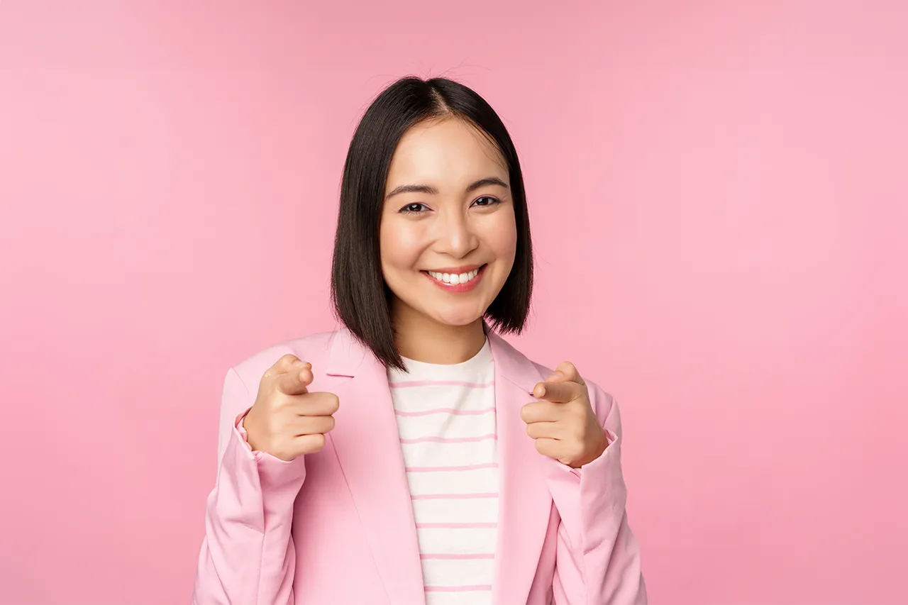 Blije Aziatische vrouw wijst met twee vingers naar de kijker met roze jasje tegen roze achtergrond